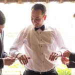 Groom in front of a window having his cufflinks fastened by two best men