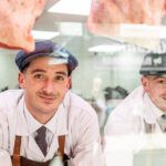 Two butchers in flat caps peering through hanging meat in the window